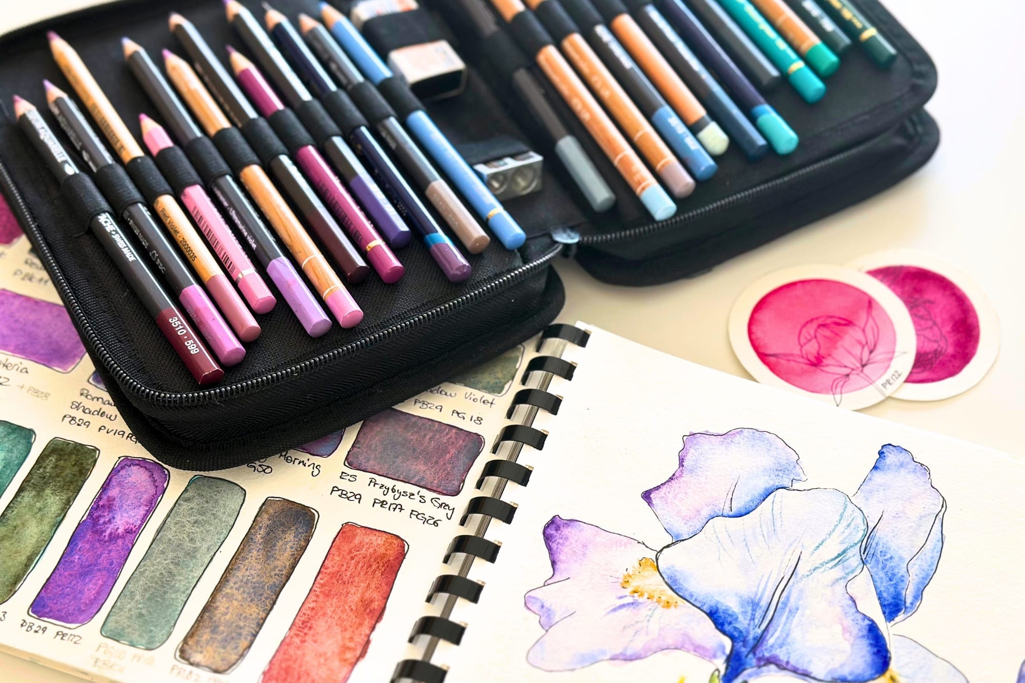 A case of watercolor pencils next to a sketchbook with an iris study